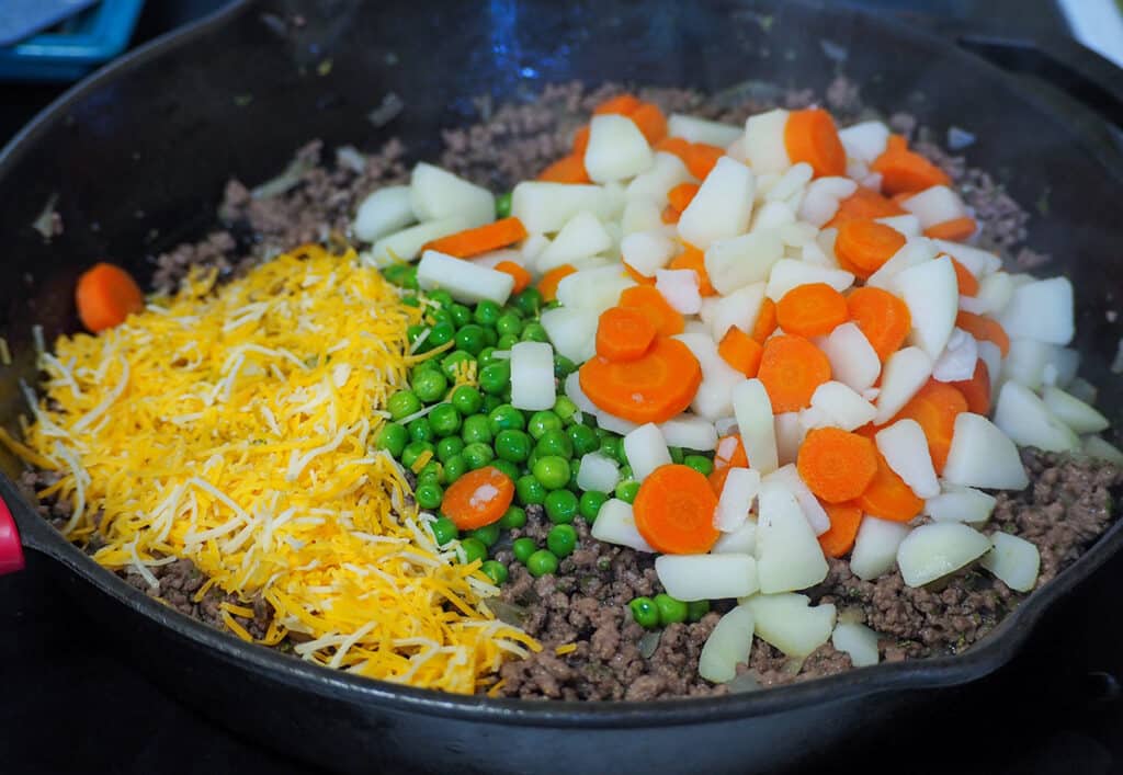 cooked ground beef in pan topped with carrots, potatoes, frozen peas, and shredded cheese waiting to be stirred together