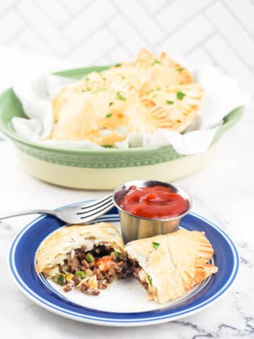 cornish beef pasty on white plate with blue rim in front of oval bowl with beef pasties