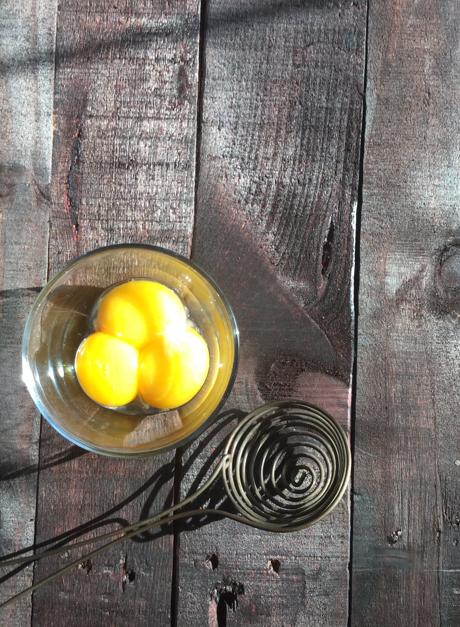 Three egg yolks in a clear bowl on a primitive wooden table with an antique whisk