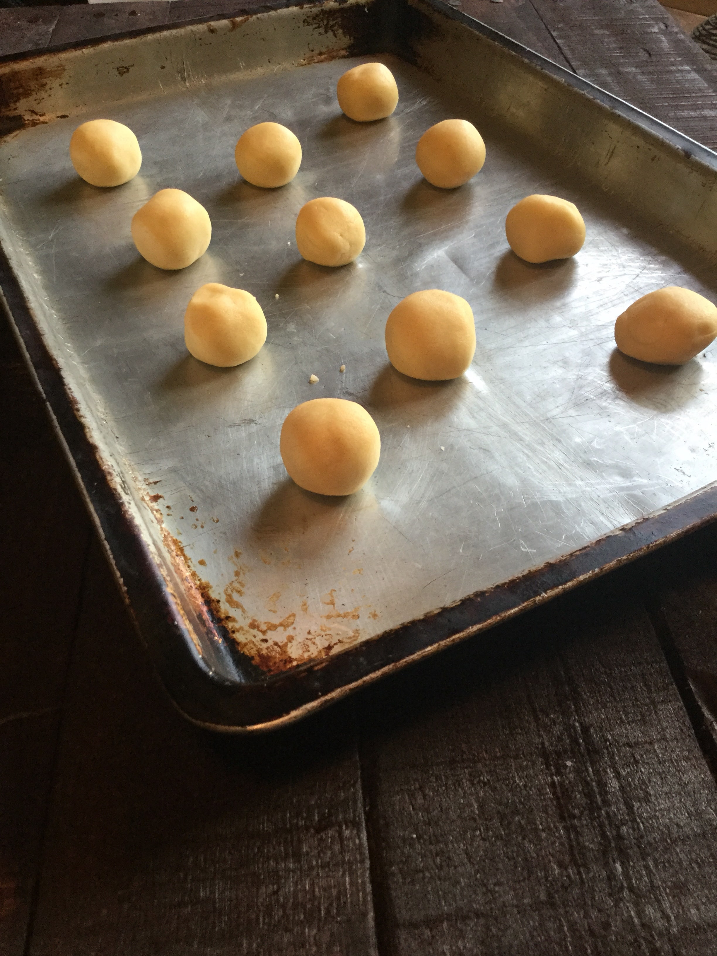 Metal baking sheet laying on a dark primitive wooden tabletop with raw cookie dough balls on sheet ready to go into the oven 
