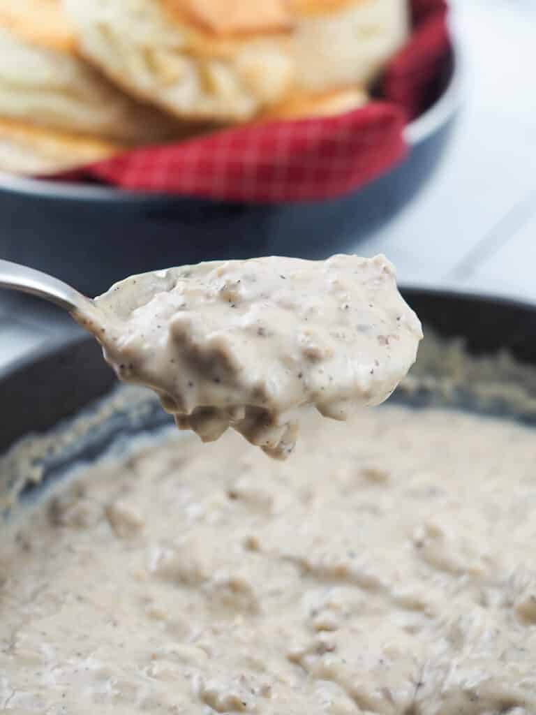 sausage gravy on suspended spoon above cast iron pan filled with gravy, biscuits in background