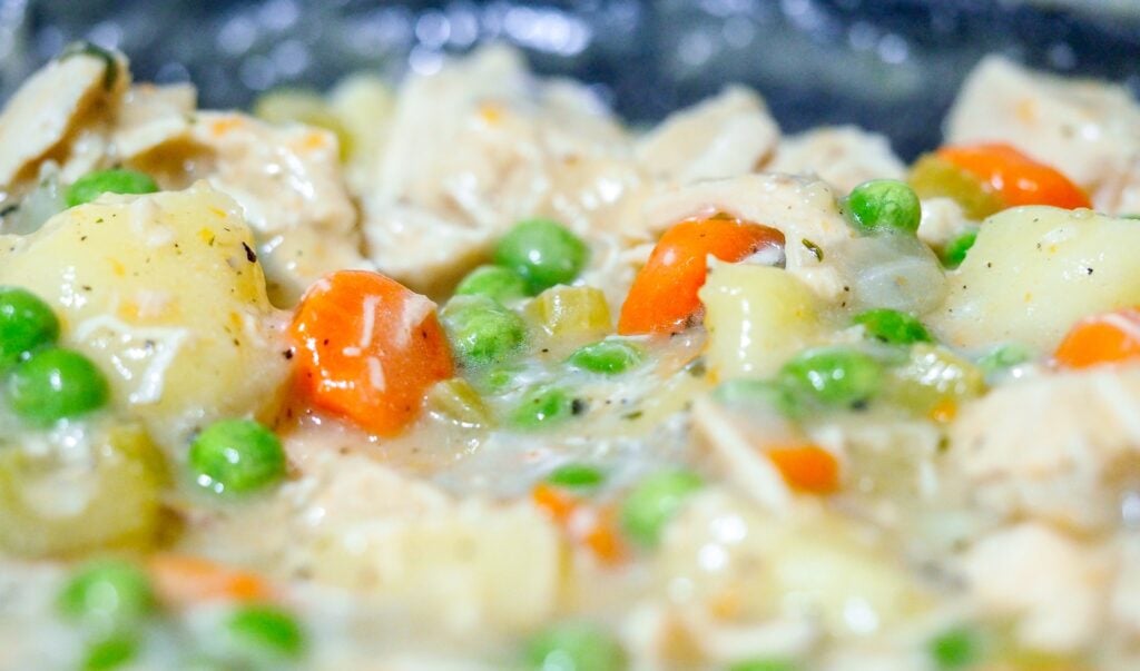 chicken, potatoes, carrots, peas, and onions in a white sauce simmering in a cast iron pan on a stove for chicken pot pie