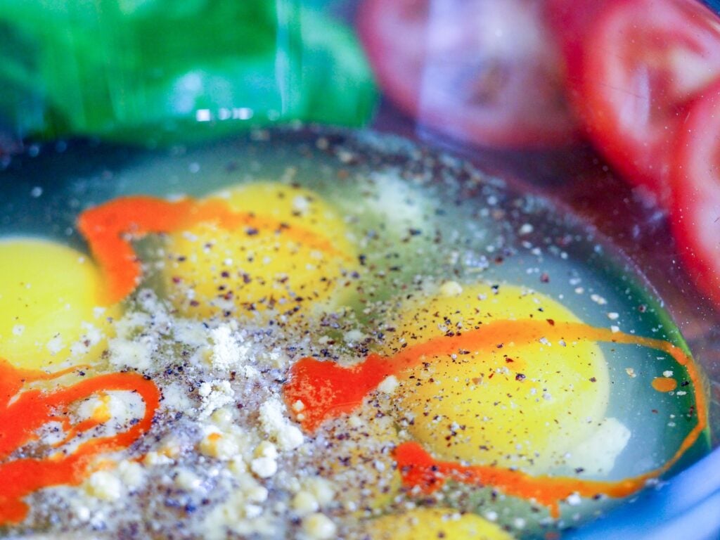 raw eggs in a green glass bowl with hot sauce and spices with vegetables in background