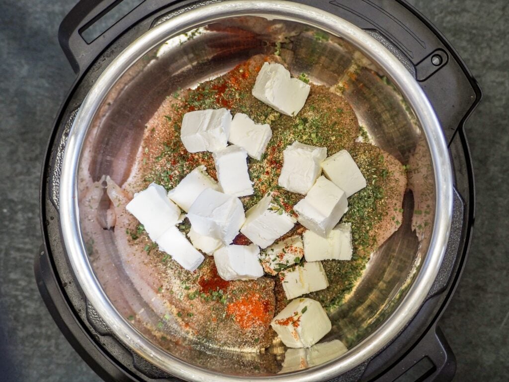 overhead view of chicken breasts in an instant pot in broth topped with spices and cream cheese