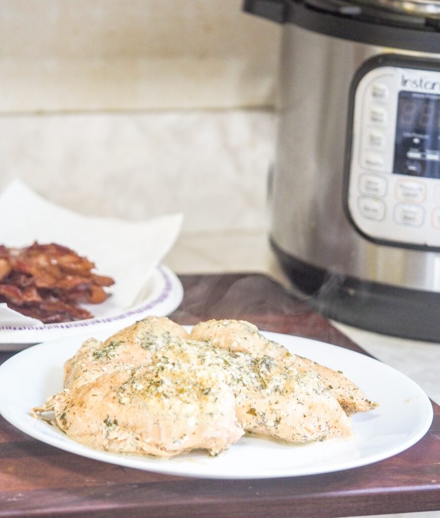 cooked seasoned chicken on white plate sitting on a mahogany cutting board with instant pot in background to the right and plate of cooked chicken to the left