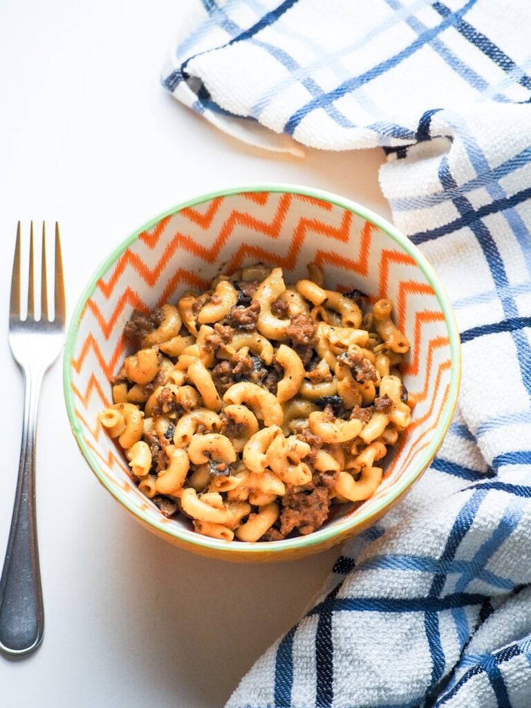 overhead shot of cheeseburger macaroni and cheese in white orange striped bowl with blue and white towel with fork to left