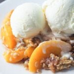 close up view of peach crisp topped with two scoops of vanilla ice cream on white plate