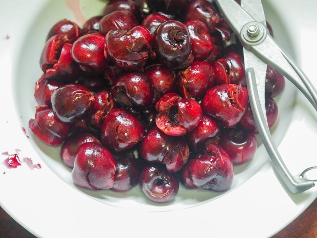 pitted cherries in a white bowl with pitting tool to the right side of the bowl
