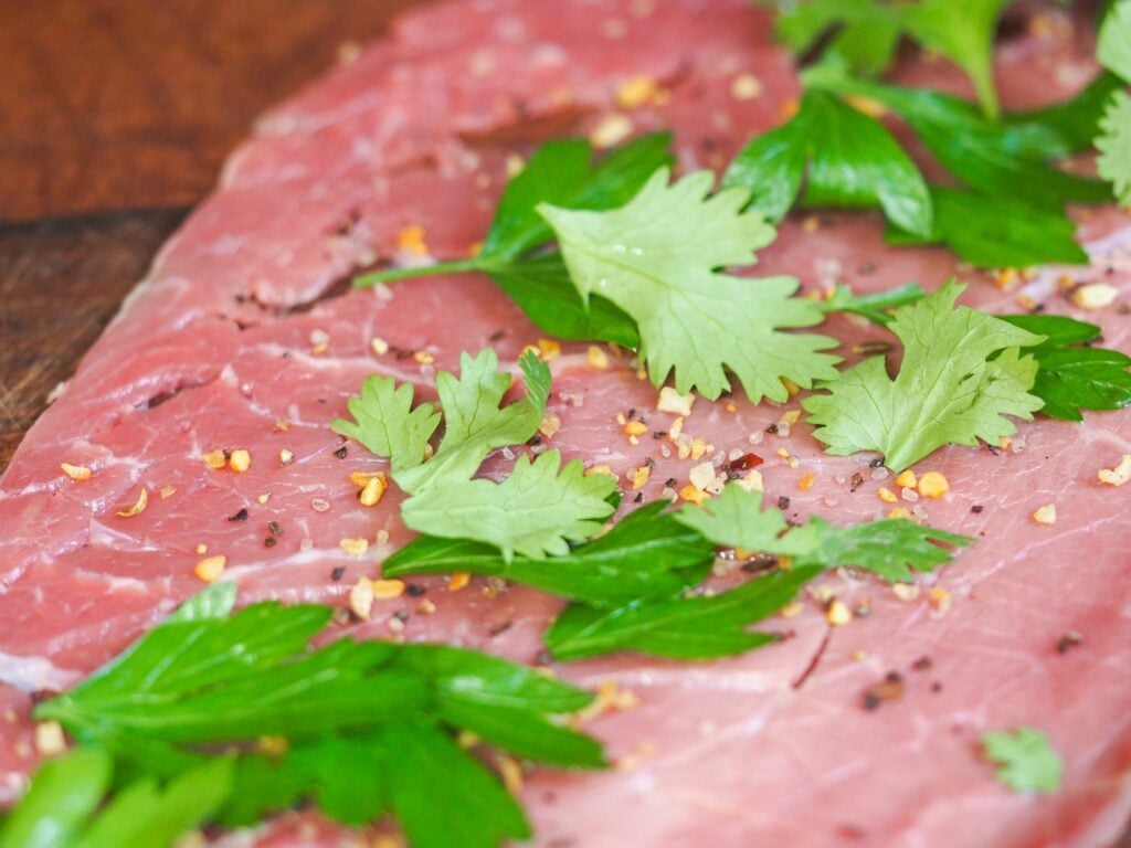 thin cut beef with seasoning and herbs laying flat on a wooden cutting board