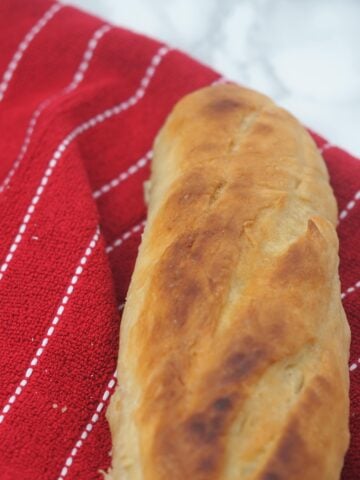 horizontal view of the top portion of a crusty french baguette sitting on a red towel with thin white stripes on a marble countertop