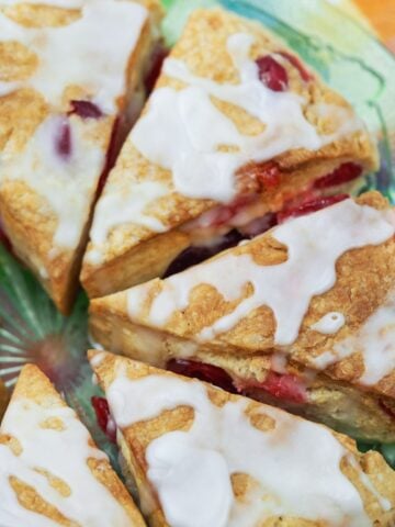 close overview of cherry scones drizzled with glaze on a clear green plate on top of multi-colored napkin in check pattern with pie server to right