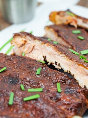 rack of ribs partially sliced on white platter sprinkled with chopped chives with bbq sauce in top left corner on wooden cutting board