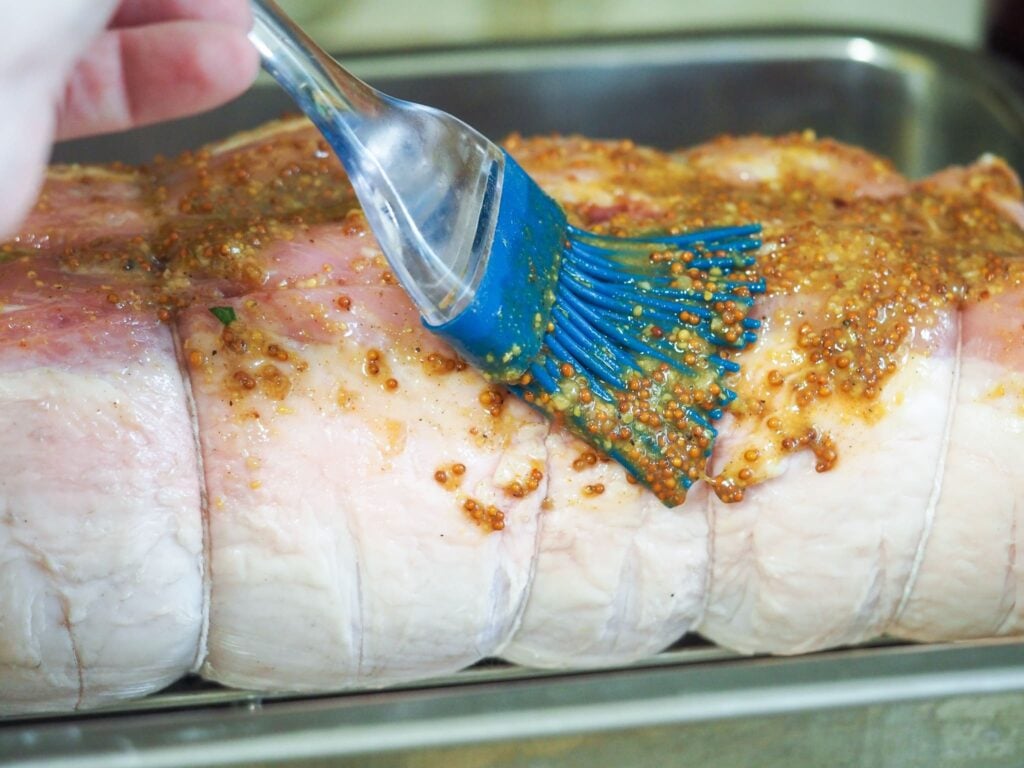 side view of tied stuffed pork loin with brush brushing on the mustard rub in metal baking pan. 