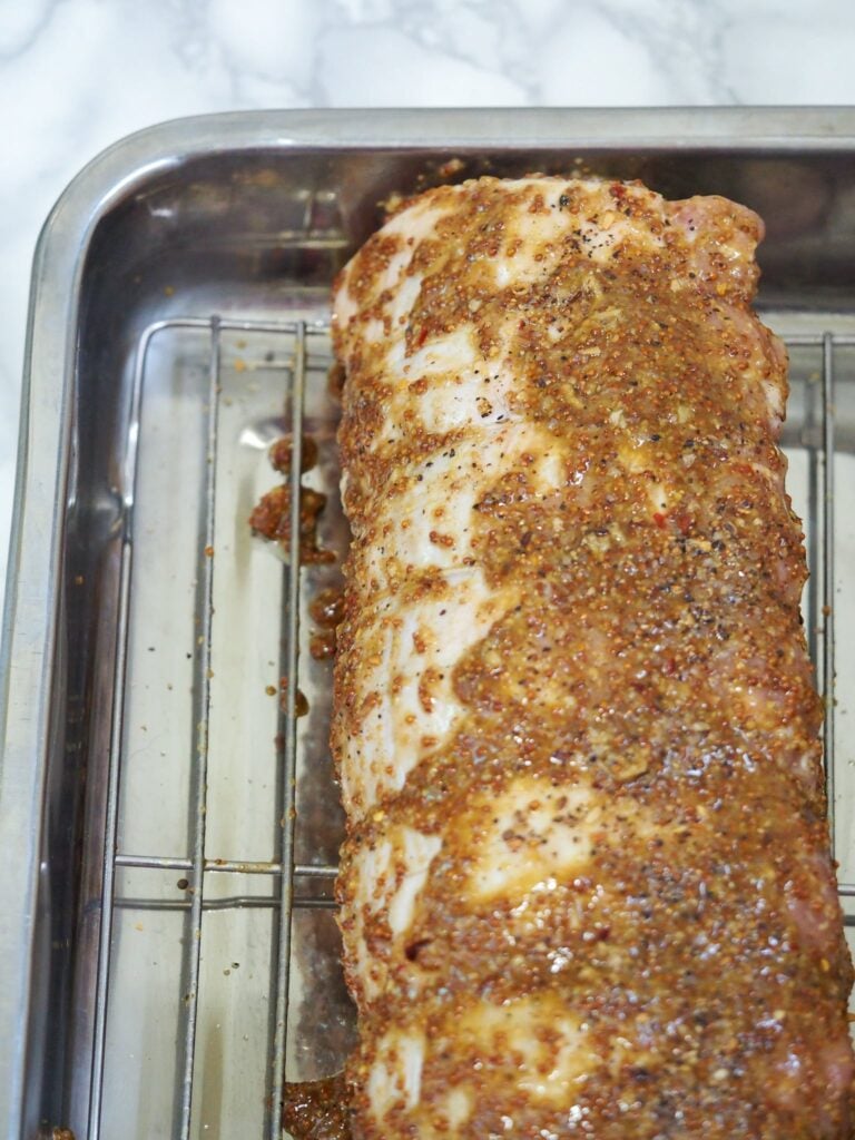 vertical image of pork loin covered in mustard rub in roasting pan on rack