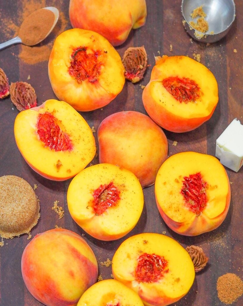 overhead shot of halved and pitted peaches on dark brown cutting board surrounded by pits, spices, butter, and brown sugar