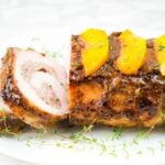 side view of stuffed pork loin stuffed with grilled peaches on white platter with fresh thyme garnish