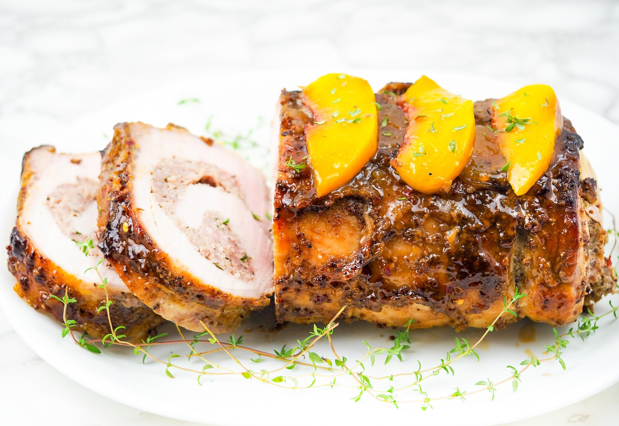 side view of stuffed pork loin stuffed with grilled peaches on white platter with fresh thyme garnish