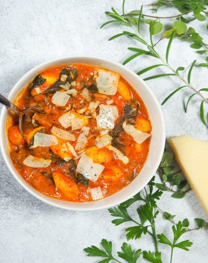 overhead view of sausage gnocchi oup in white bowl on concrete background with wedge of Parmesan cheese and fresh herbs to the lef