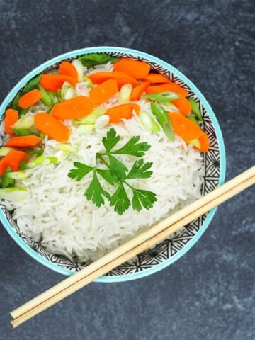 bowl of coconut rice on black textured background in bowl with blue edge garnished with sliced carrots and green onions with parsley, chop sticks resting on bottom right of bowl