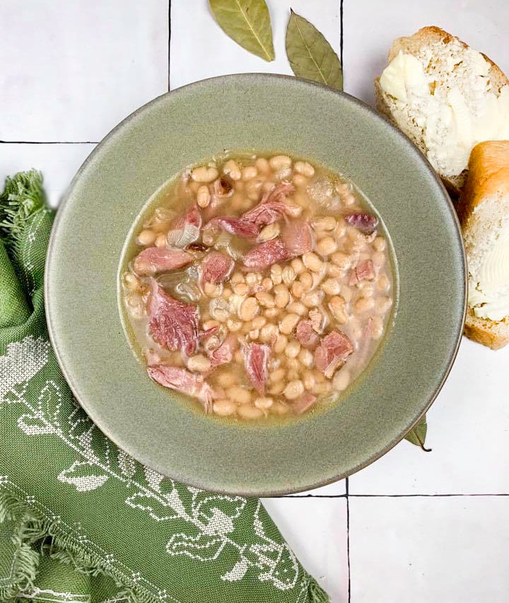 overhead view of navy bean soup in olive green bowl with green napkin at bottom left, sliced and buttered French bread at top right, on a tile surface with bay leaves scattered