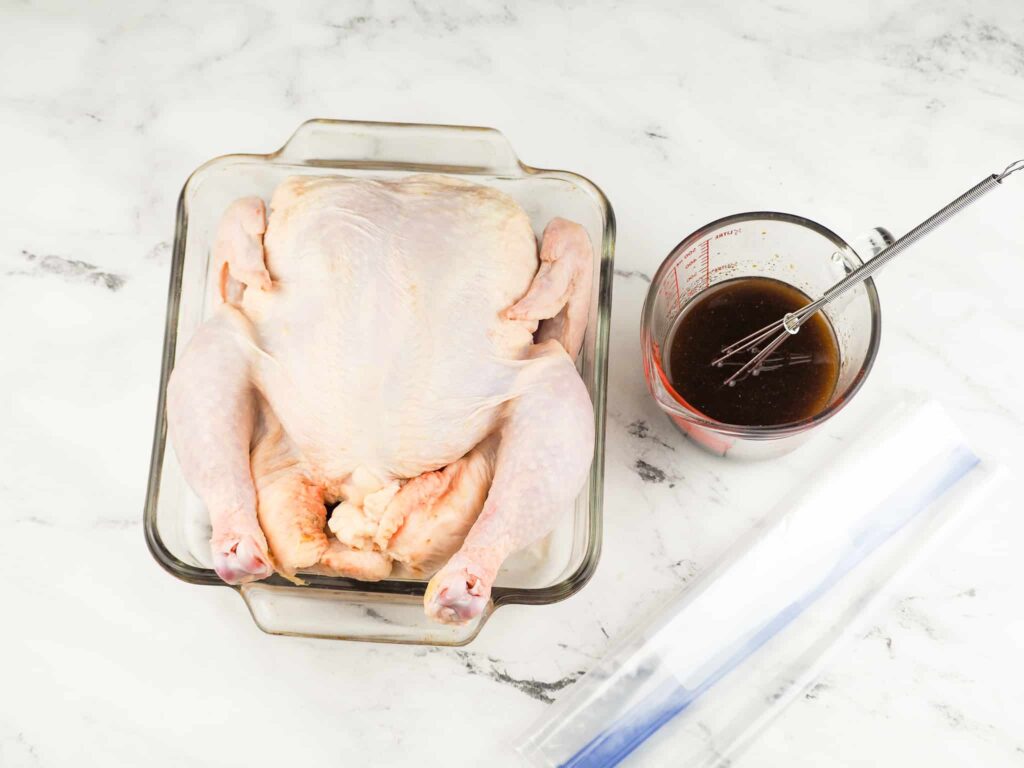 overhead view of whole chicken in small square clear glass baking pan with marinade in measuring cup with plastic bag