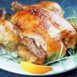 roasted whole chicken on white platter surround by orange quarters and fresh rosemary on black background
