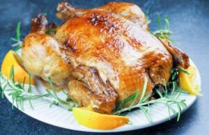 roasted whole chicken on white platter surround by orange quarters and fresh rosemary on black background