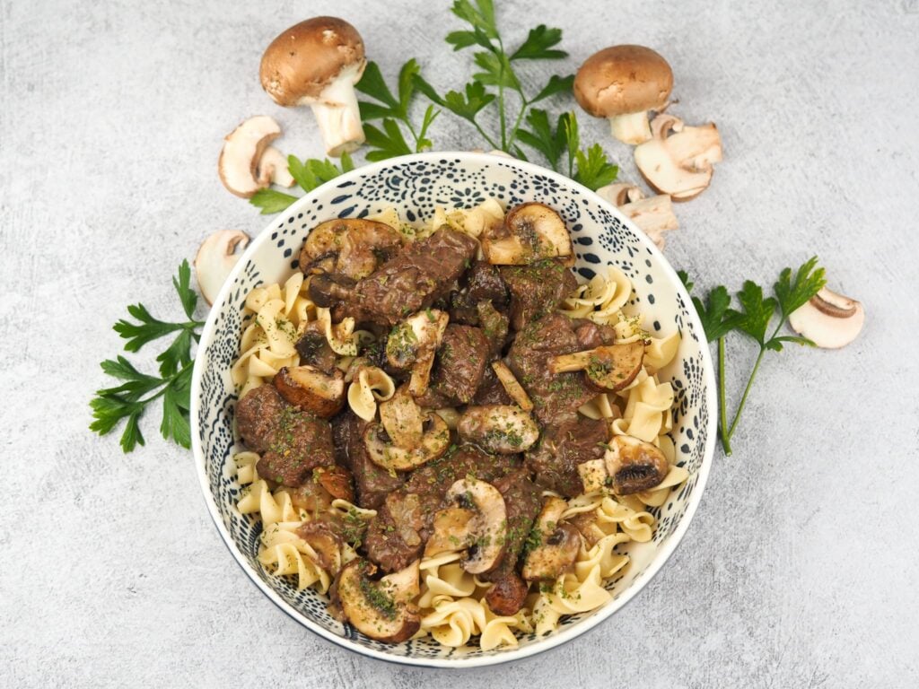 bowl of beef tips over egg noodles on grey concrete background circled with fresh mushrooms and parsley