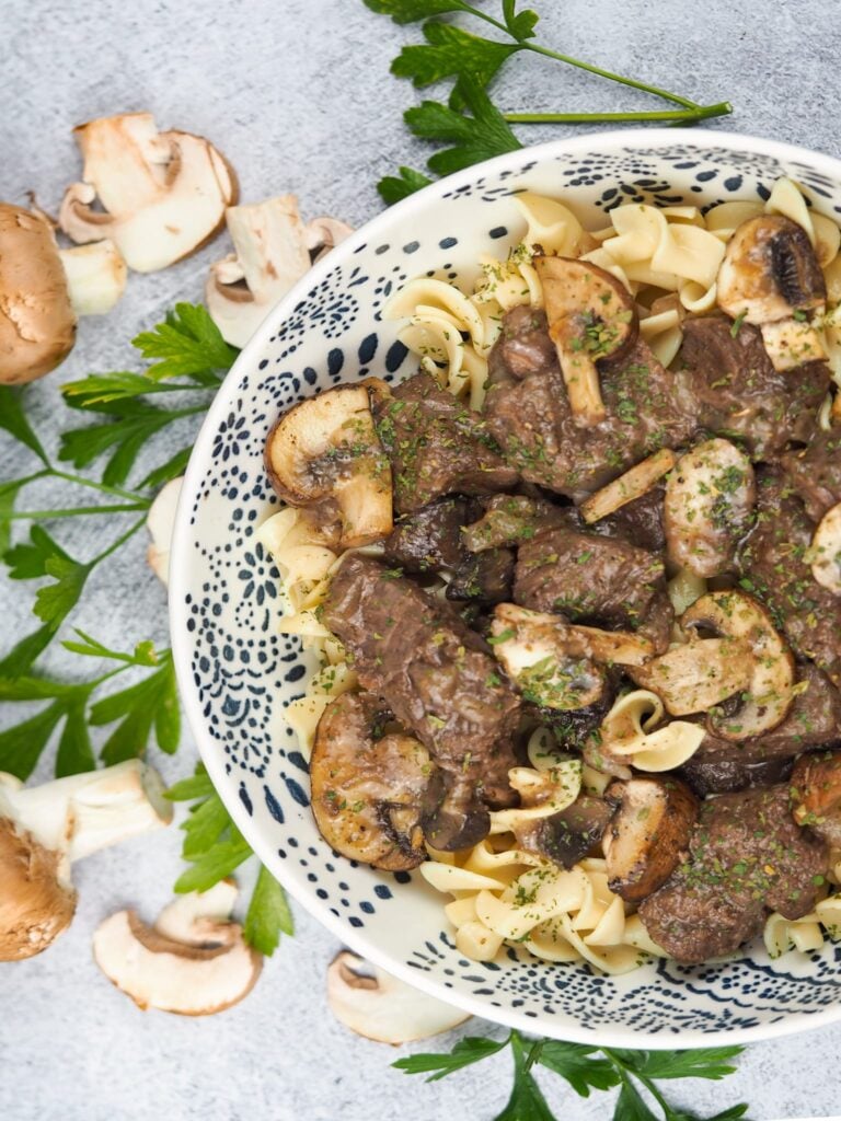 overhead view of beef tips with mushrooms over egg noodles in white bowl with blue graphic designs on concrete background with fresh herbs and mushrooms