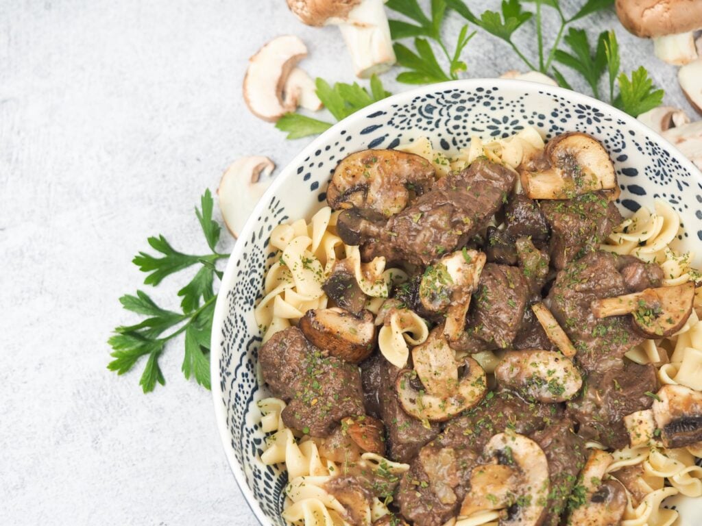 sirloin tips with mushroom gravy in white ceramic bowl with dark blue flowers on concrete background with fresh parsley and whole or sliced mushrooms