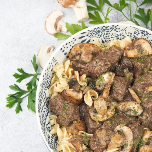 sirloin tips with mushroom gravy in white ceramic bowl with dark blue flowers on concrete background with fresh parsley and whole or sliced mushrooms