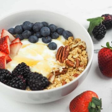 yogurt in white bowl on granite counter top next to berries topped with honey, granola, and berries