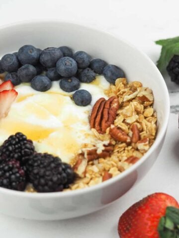 yogurt in white bowl on granite counter top next to berries topped with honey, granola, and berries
