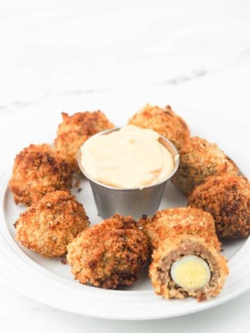 vertical picture of scotch eggs on white plate circling dipping sauce in metal cup