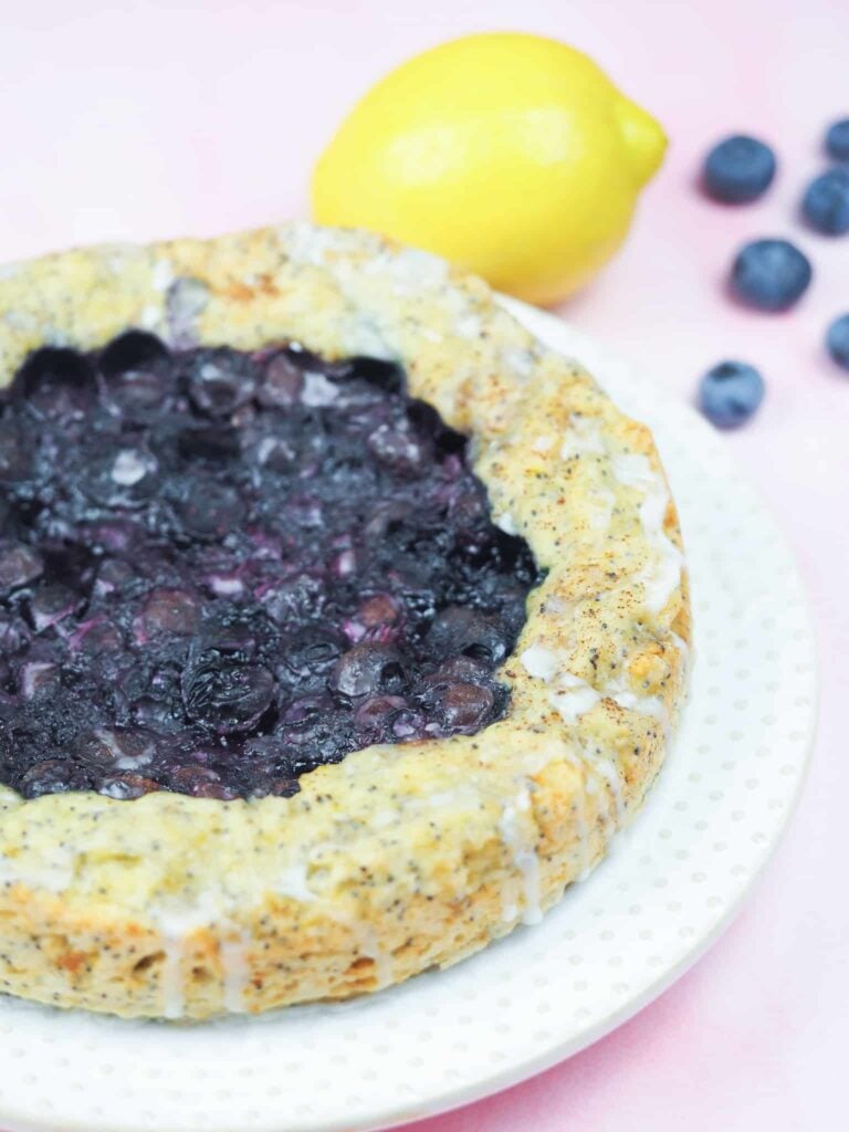 blueberry poppy seed cake on white plate offset to bottom left with lemon and blueberries on top left on pink marble background