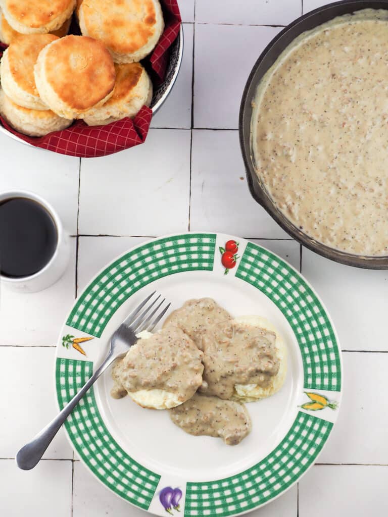 biscuits and gravy on plate topped with sausage gravy with bowl of biscuits in top left and pan of gravy in top right