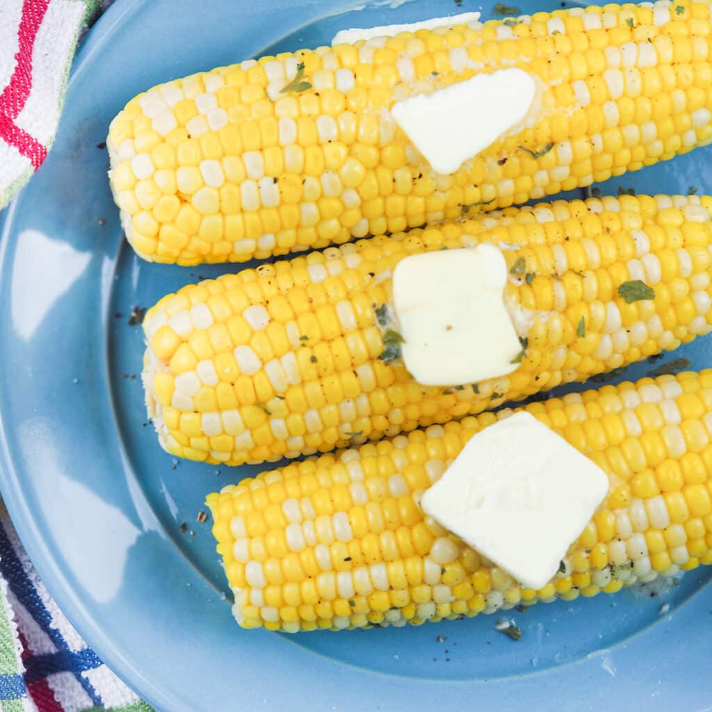 corn on the cob topped with butter pats on blue plate surrounded by multi colored towel