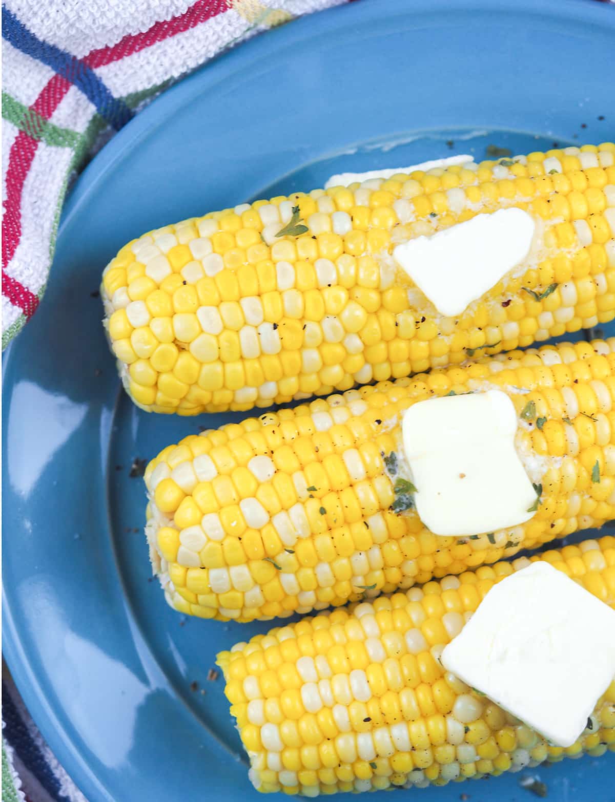 close  up view of three ears of corn on the cob on blue plate with butter pats and sprinkled parsley