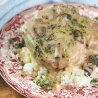 Bone-in pork chop on bed or rice topped with mushroom gravy and parsley on red and white vintage plate.