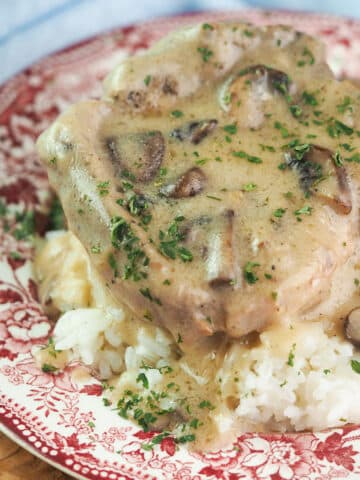 Bone-in pork chop on bed or rice topped with mushroom gravy and parsley on red and white vintage plate.