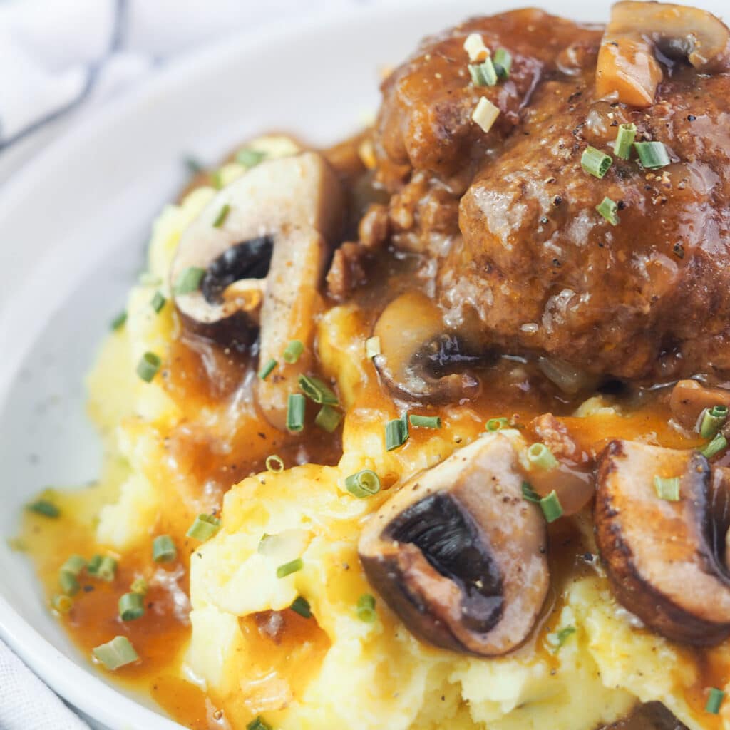 Salisbury steak on top of mashed potatoes with mushroom gravy and chives on white plate.