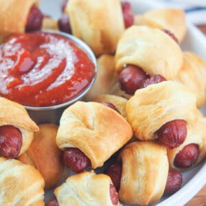 Close up view of mini pigs in a blanket on white plate with spicy ketchup dipping sauce.