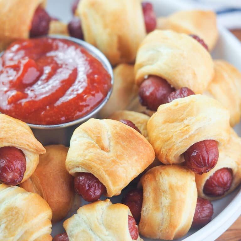 Close up view of mini pigs in a blanket on white plate with spicy ketchup dipping sauce.
