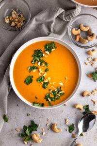 Cashew carrot ginger soup in white bowl on concrete background with fresh herbs and cashews.