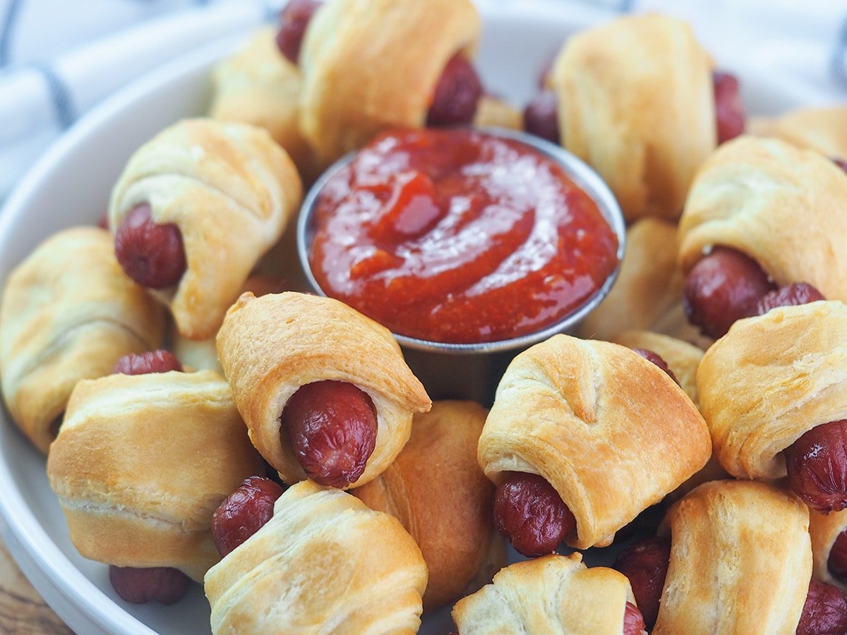 Pigs in a blanket on white serving platter arranged around ketchup in a small cup.